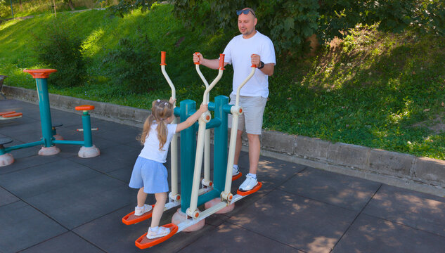Active Father And Young Daughter Exercising Together On Double Outdoor Elliptical Fitness Equipment In Park
