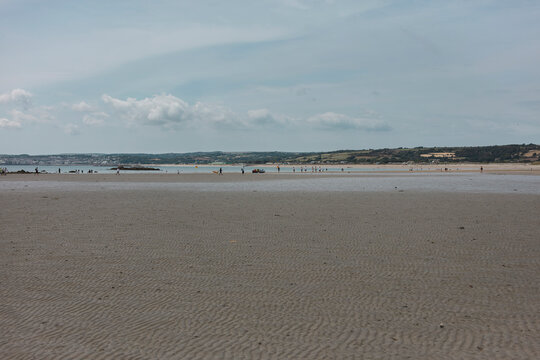 Wide rippled sand flats at low tide in Mount's Bay, Cornwall, with tiny figures at the waterline and green hills across the bay.