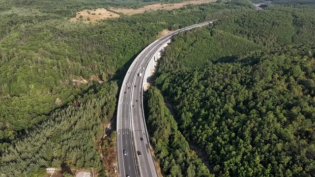 Hyperlapse aerial view of flowing traffic on a curved highway viaduct passing through a dense green forest during summer.