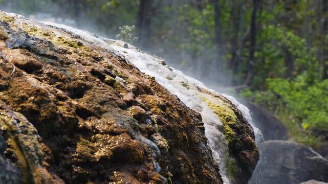 Extremely hot geothermal spring water flows over mineral-covered rocks in a mountain forest, releasing steam and a characteristic hydrogen sulfide odor from the naturally heated mineral source.
