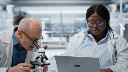 Pathologist examines a biopsy slide while a coworker records diagnostic findings on a laptop, working together to identify disease markers in a clinical lab. © DC Studio