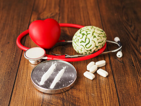 Stethoscope rests beside brain model and tow line of drugs on small mirror on wooden surface. Medical equipment and brain suggest focus on mental health damage due to narcotic use.