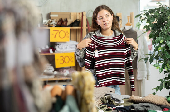 Young female shopper choosing knitted sweater in clothing store