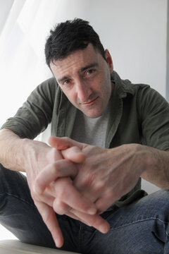 Close-up portrait of a mid-adult Latino man leaning forward with hands clasped, looking directly into the camera with a subtle smirk and an engaging, skeptical expression.