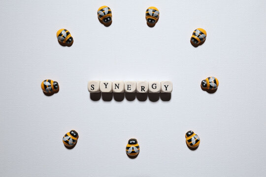 Bee figures surround wooden letters spelling &ldquo;SYNERGY&rdquo; in a flat lay concept representing teamwork, collaboration and collective success.