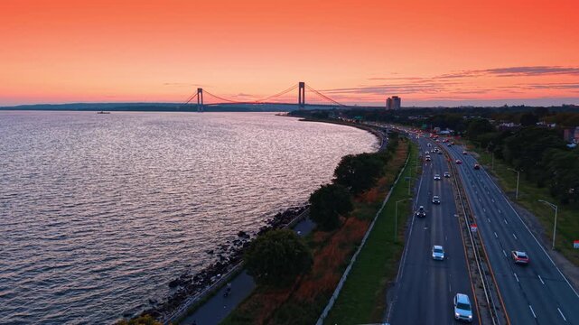 Lively traffic by the waterfront along the waterscape at sunset time. View on the Verrazzano-Narrows Bridge against orange sky in the backdrop.