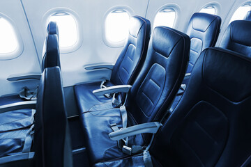 Obraz na płótnie Canvas Empty blue passenger seats line a commercial airplane cabin beside bright windows. Seat belts armrests and leather upholstery fill the aircraft interior for passenger travel background.