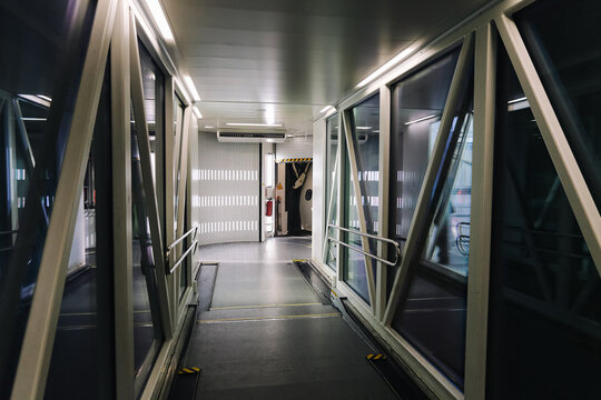 Enclosed passenger boarding bridge corridor leads toward an aircraft door. Glass panels metal beams handrails and overhead lights line the narrow airport walkway before a flight departure background.