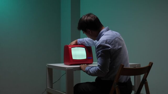 Man sits at a dim table intently adjusting a vintage red tv with a flickering screen, conveying frustration with outdated technology and the struggle to connect in a digital age