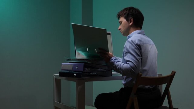 Exhausted office worker sitting at a desk in a dark room and searching for files in a ring binder late at night, demonstrating burnout, stress, and overwhelming paperwork in a corporate job