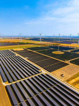 High angle view of solar energy farm project. Large solar panel field with wind turbines on background landscape, bright clear weather.