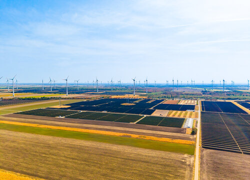 Panoramic aerial view of solar farm and wind turbines. Photovoltaic arrays and white generators cover yellow agricultural fields under clear sky.