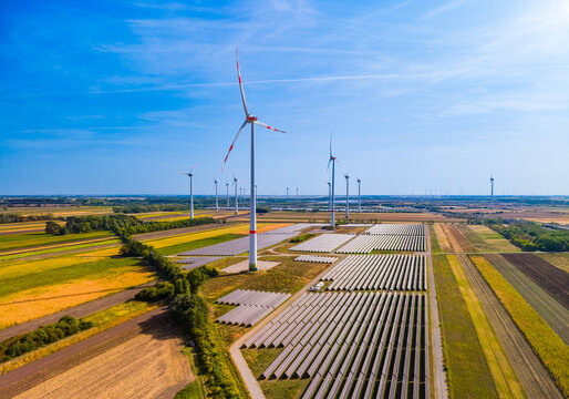 Aerial view of wind turbines and solar power plant on rural fields. Patterned crop plots surround white generators and rows of light reflecting panels.