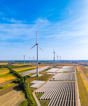Aerial shot of tall wind turbine above solar panels. A large white generator rises over ground mounted photovoltaic collector rows.