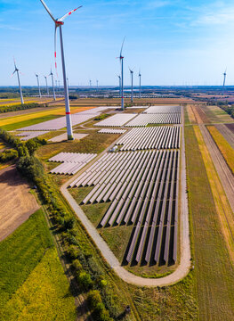 Solar panel plant, photovoltaic field layout near wind turbines. High angle vertical shot of solar panel rows installed on ground beside tall white wind turbine