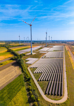 High wind turbine, green energy farm with solar panels. Vertical aerial perspective of tall wind turbine rising above solar panel rows and farm fields