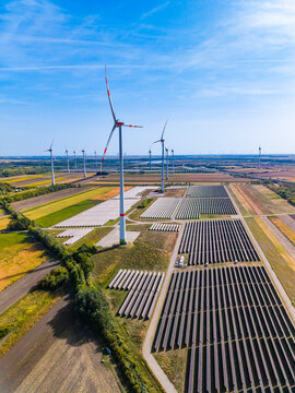 Solar panel park with wind turbines in rural field. High angle view of photovoltaic power station, white wind generators, blue clear sky, green and yellow agricultural fields.