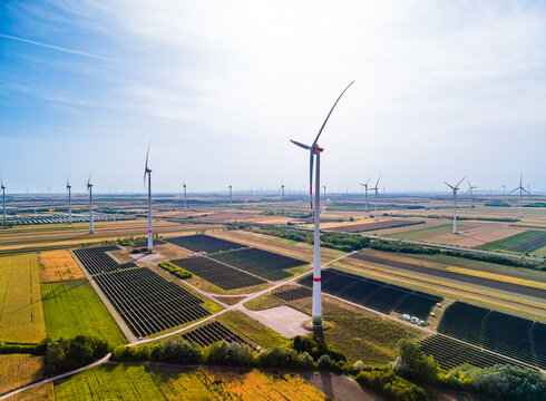 High angle view of solar panel field with wind turbines. Sunlight illuminates structured photovoltaic arrays positioned beside cultivated crops on a clear sunny day.