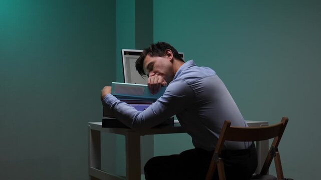 Young businessman experiencing exhaustion and stress, sleeping with his head resting on a stack of binders while working late at night in a dark office
