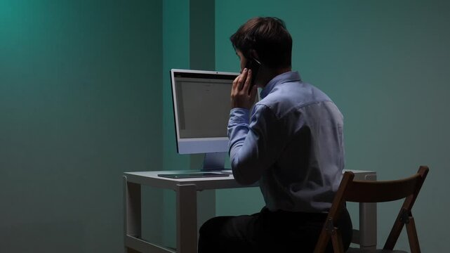 Young man sitting at a desk, talking on a smartphone while looking at a computer monitor, focusing on remote work and business communication in a modern office