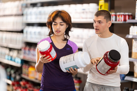 Selective young guy and girl choosing protein powder jar in large sports nutrition store