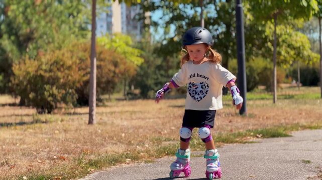 Little girl wearing a protective helmet and knee pads confidently roller skates along a park path. Active recreation, sports, childhood and outdoor lifestyle concept