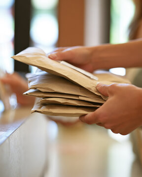 Paper parcel stack handed over at postal counter, showing adult hands, sealed envelopes, warm indoor light, and realistic delivery service
