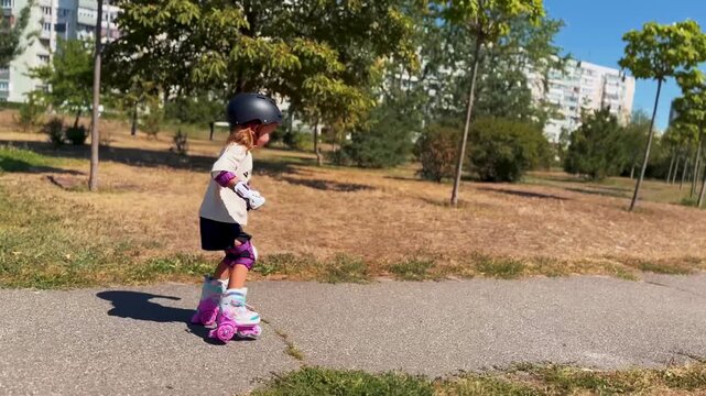 Little girl wearing a helmet and protective gear roller skating along a park path. Children's sports, active recreation, movement and happy carefree childhood concept