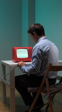 Vertical video man in a collared shirt sitting at a table, intensely looking at a red vintage television with a blank screen, suggesting themes of introspection, media consumption, and nostalgia