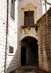 A stone archway connects traditional limestone buildings above a shaded pedestrian passage featuring decorative window pediments and wooden shutters in the Old Town of Kotor, Montenegro © Gregory
