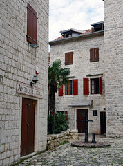 A cast iron water pump stands in a cobblestone square framed by traditional limestone buildings with wooden window shutters and an antique store entrance in Kotor, Montenegro © Gregory