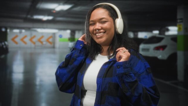 Young woman wearing headphones holding them to her ears as she smiles in a parking building, blue flannel and white top; joy.