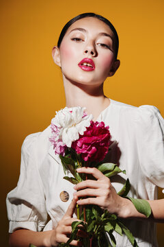 Fashion model with glossy makeup poses holding red and white flowers. Styled with sleek hair and soft lighting on warm orange background. Creative beauty and fashion concept.