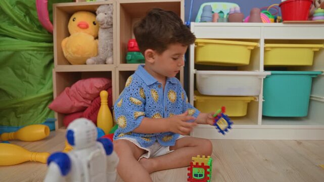 Boy sitting on the floor assembling interlocking plastic blocks and reaching with his hands toward a toy robot in a playroom; concentration.