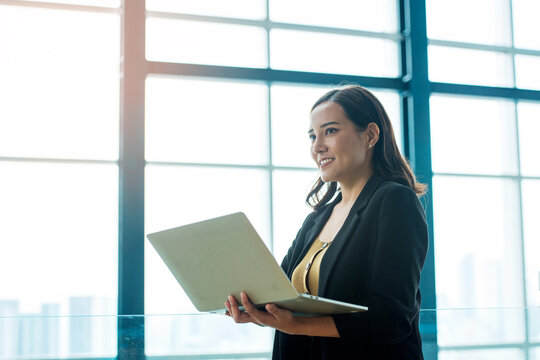 Professional Asian Businesswoman Using Laptop in Modern Office with Large Windows