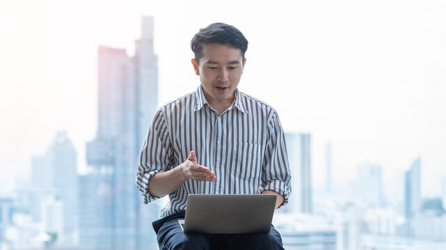 Asian Businessman Using Laptop in Modern City Office with Urban Skyline Background
