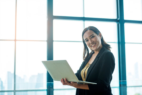 Professional Asian Businesswoman Using Laptop in Modern Office with Large Windows