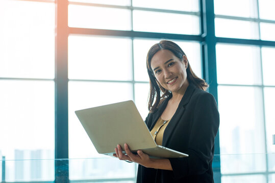 Professional Asian Businesswoman Using Laptop in Modern Office with Large Windows