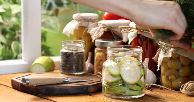 Pickled products. Woman putting dill into glass jar with zucchini slices at wooden table, closeup