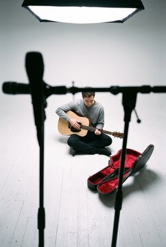 Young male musician sitting cross-legged on floor playing acoustic guitar during recording session in white photo studio
