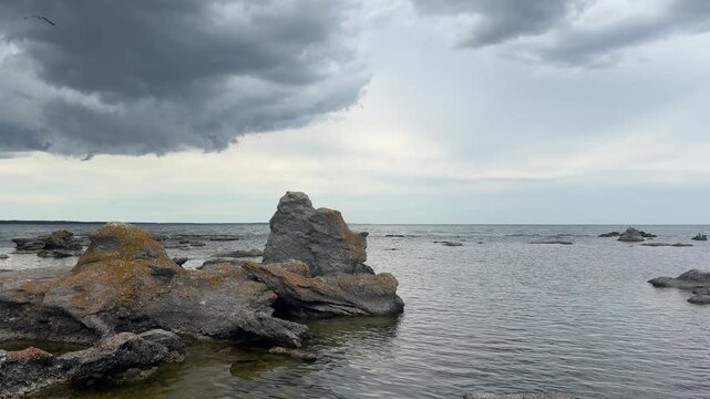 Wide cinematic 4K pan across ancient limestone rauk monoliths standing in the calm Baltic Sea with seabirds flying under a moody sky in Gotland, Sweden.