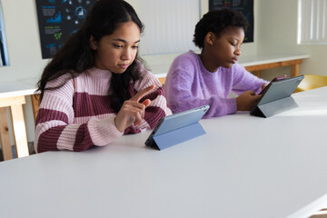 African American teen girls sitting in pink and lilac sweaters tapping tablets with phone in class © WavebreakMediaMicro