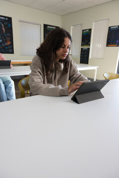 Female teen classmates sitting at class front table leaning over tablet in folio case with headset
