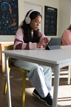 Female youth students sitting in class, tapping tablet in folio and wearing headphones