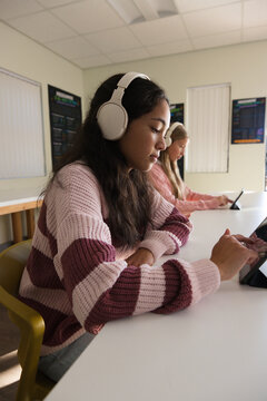 Teen female students using tablets at long white classroom table, wearing headphones and sweaters