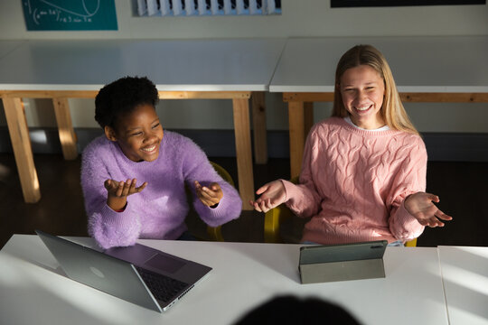 Two female teens laughing, gesturing in class in lavender, pink sweaters using laptop, folio tablet