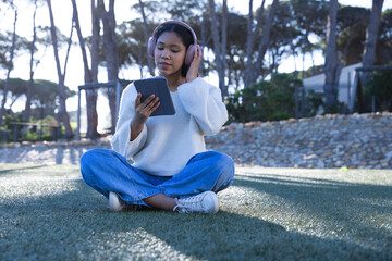 Female child sitting cross-legged on turf in park holding tablet and adjusting pink headphones © WavebreakMediaMicro