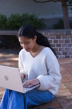 Asian female teenager wearing white sweater, blue jeans, typing on silver laptop at campus plaza