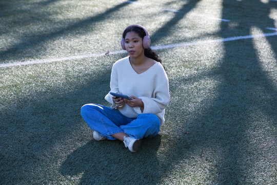 Asian girl child sitting crosslegged on turf field holding tablet wearing sweater, pink headphones