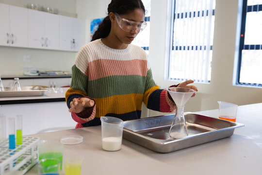 Female child wearing goggles and sweater, positioning funnel over lab flask on tray in school lab